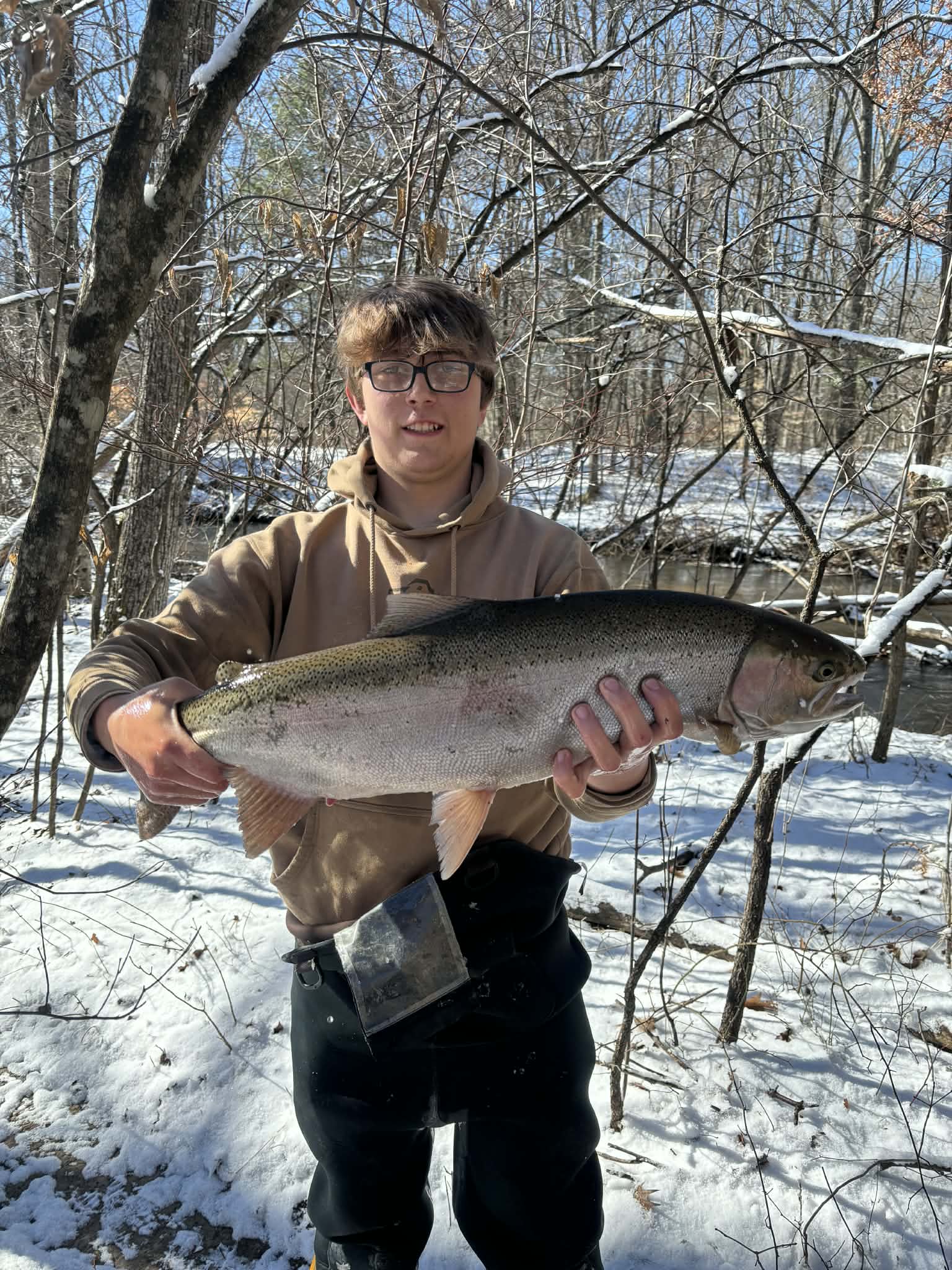 Liam, wearing glasses and a brown top, holding a fish beside the river